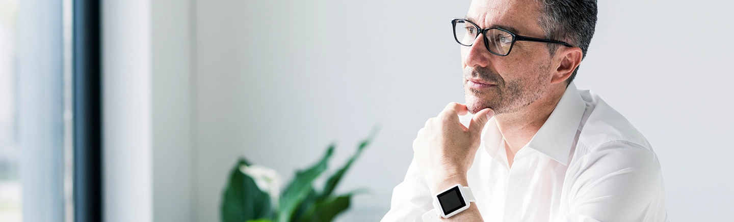 Person in a bright home office wearing a white shirt and smartwatch, seated near a window with a green plant in the background, illustrating how to maintain wellbeing while working from home.