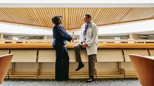Two professionals standing and speaking inside a large, contemporary public building with open seating and wood‑panelled ceilings.